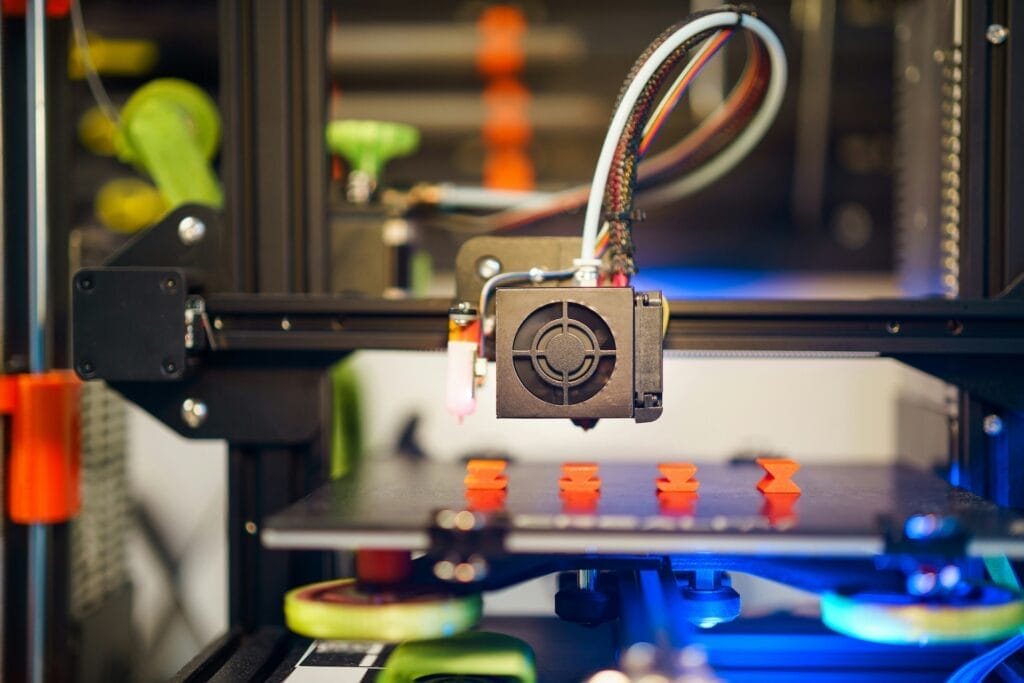 A 3D printer in operation, producing several small, orange objects on its build platform.