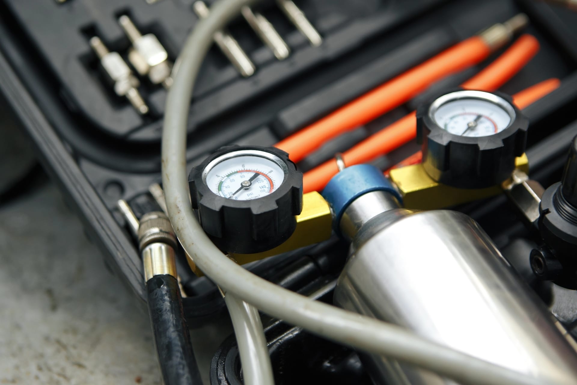 Close-up of a pressure gauge device with two dials and connecting hoses, placed inside a toolbox containing various tools and cables.