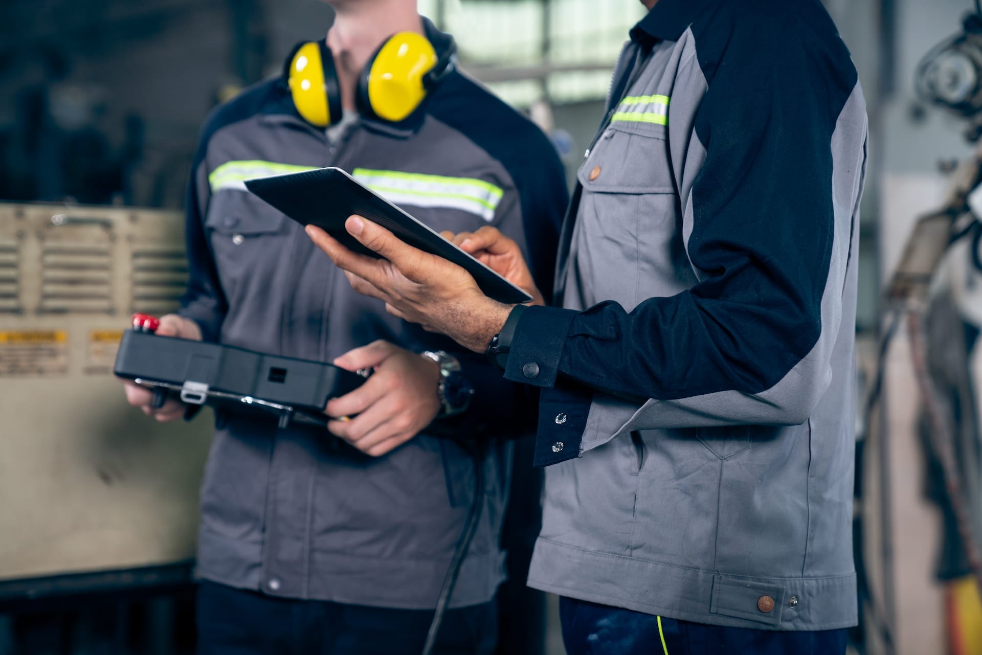 Two factory workers in uniform stand in an industrial setting, one holding a control box and the other using a tablet. Both are focused on their tasks.