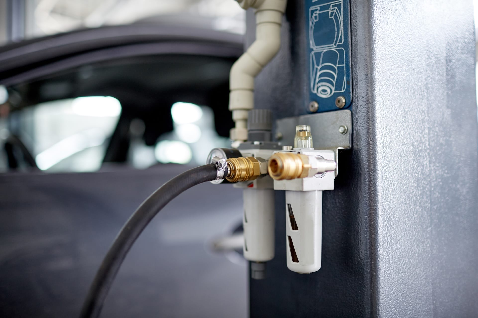 Close-up of equipment at a car service station. The image shows two cylindrical filters connected to hoses, with a vehicle in the background.
